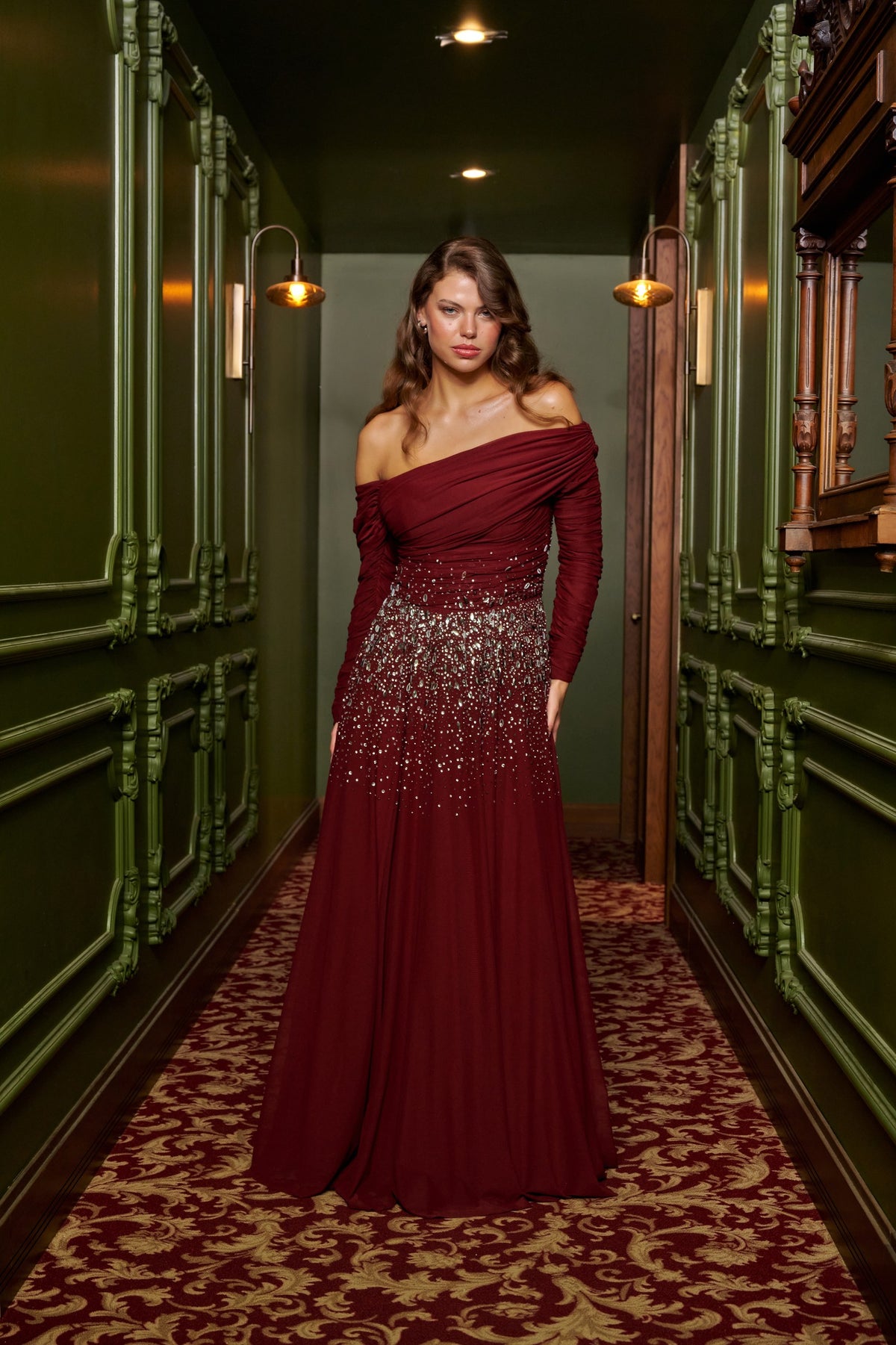 Ruby Drape burgundy off-the-shoulder evening dress with long sleeves, crystal waist embellishments and a flowing floor-length skirt.