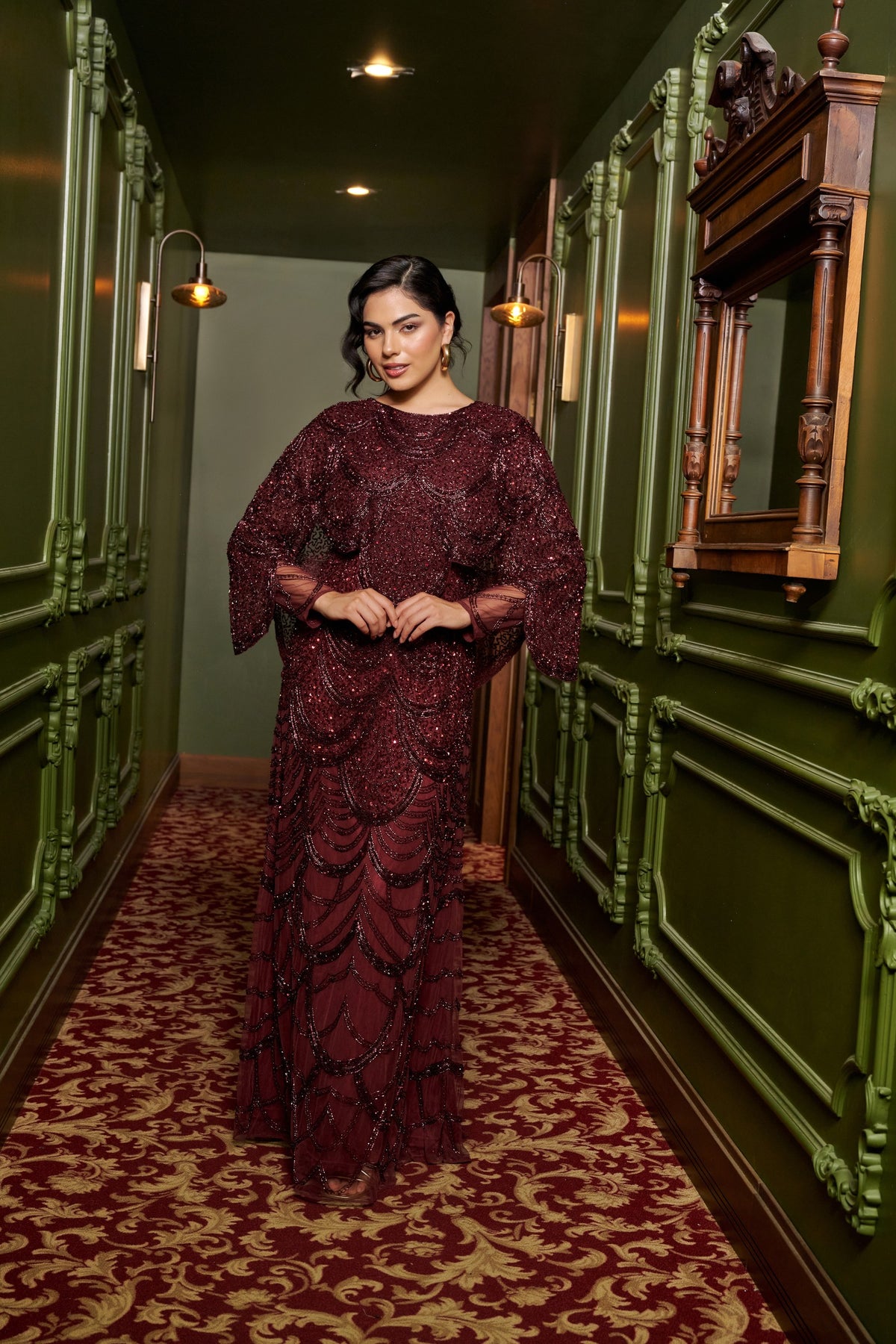 Ruby Grace burgundy embellished evening gown with a sheer cape overlay, flowing sleeves and a floor-length fitted silhouette.