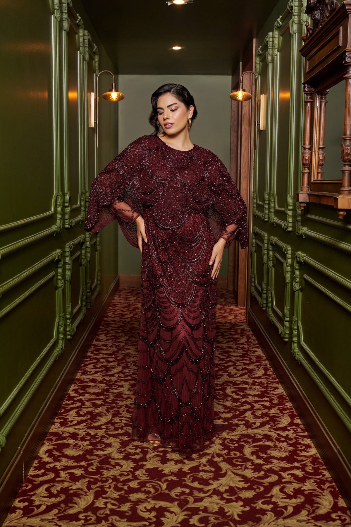 Ruby Grace burgundy embellished evening gown with a sheer cape overlay, flowing sleeves and a floor-length fitted silhouette.