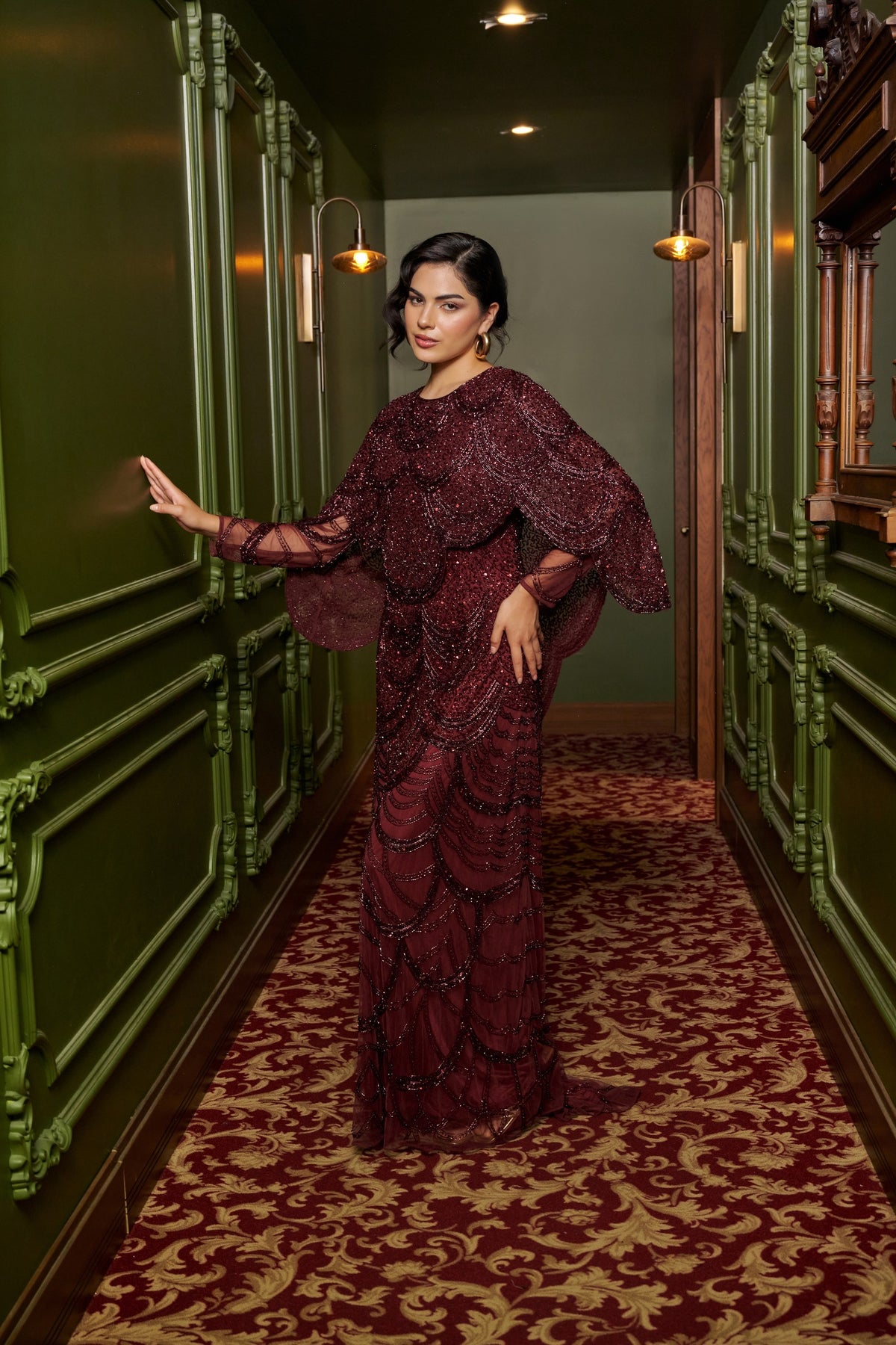 Ruby Grace burgundy embellished evening gown with a sheer cape overlay, flowing sleeves and a floor-length fitted silhouette.